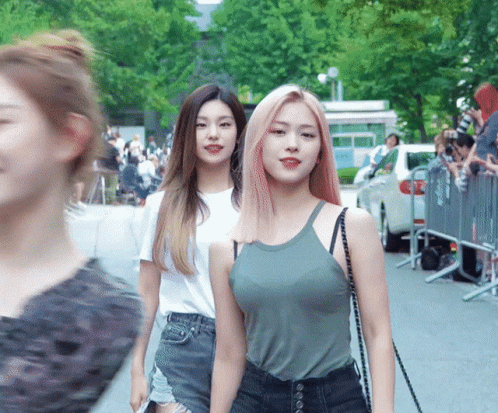 Two Women Are Walking Down A Street With A Crowd Behind Them GIF
