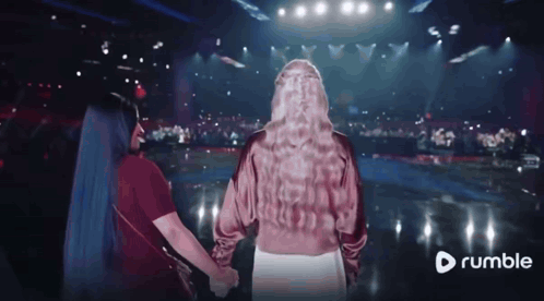 Two Women Holding Hands In Front Of A Stage With A Rumble Logo In The Corner GIF