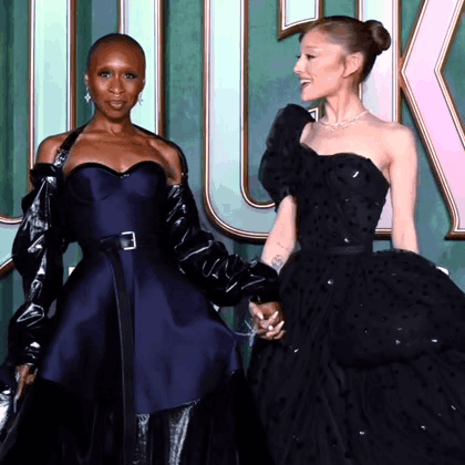 Two Women In Black Dresses Are Holding Hands And Smiling GIF