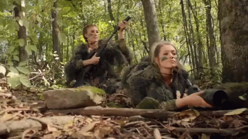 Two Women In Camouflage Are Laying In The Woods Holding Guns . GIF