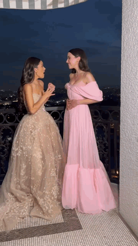Two Women In Long Dresses Are Standing Next To Each Other On A Balcony At Night GIF