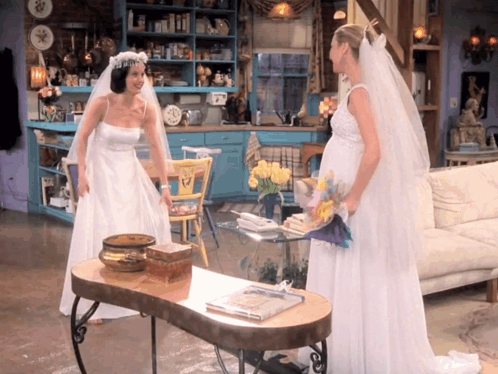 Two Women In Wedding Dresses Standing Next To Each Other In A Living Room GIF