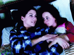 Two Women Laying On A Bed Looking At Each Other With One Wearing A Plaid Shirt GIF