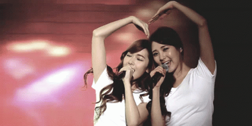 Two Women Singing Into Microphones And Making A Heart Shape GIF