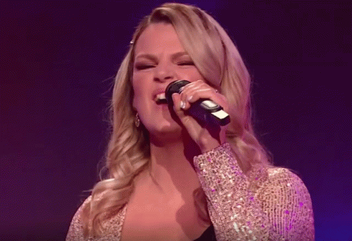 Two Women Singing Into Microphones On A Stage In Front Of A Band GIF