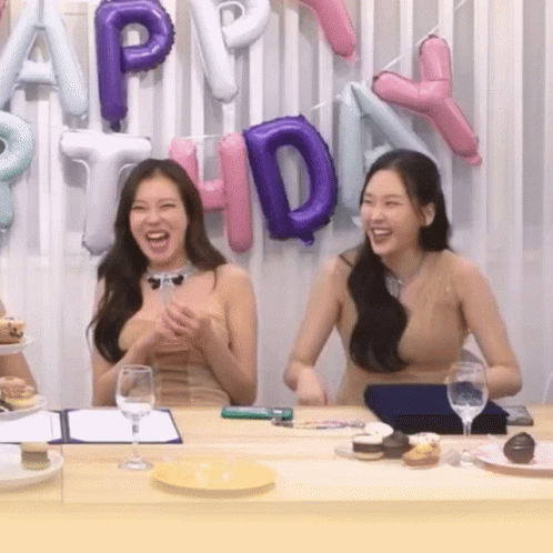 Two Women Sit At A Table In Front Of Balloons That Spell Out Happy Birthday GIF