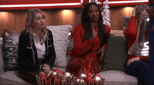 Two Women Sit On A Couch With A Bowl Of Christmas Ornaments In Front Of Them GIF