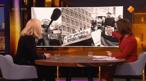 Two Women Sitting At A Table In Front Of A Screen That Says Nos On It GIF