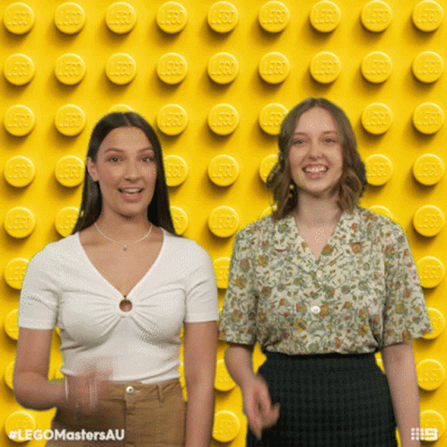 Two Women Standing in Front of Yellow Lego Wall