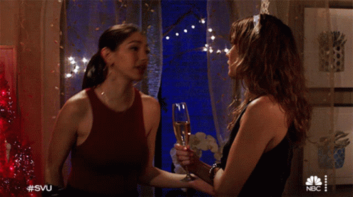 Two Women Standing Next To Each Other Holding A Glass Of Champagne With The Nbc Logo On The Bottom GIF
