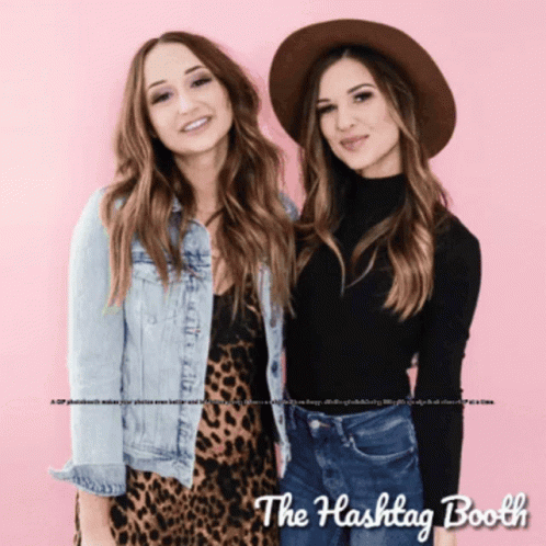Two Women Standing Next To Each Other In Front Of A Pink Background With The Hashtag Booth Written On It GIF
