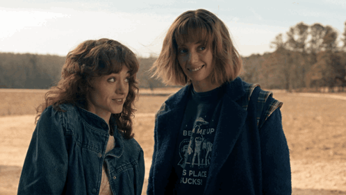 Two Women Standing Next To Each Other One Wearing A Shirt That Says Be Me Up GIF