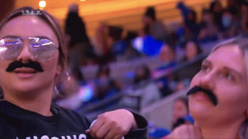 Two Women Wearing Fake Mustaches And Sunglasses Are Standing In A Crowd .. GIF