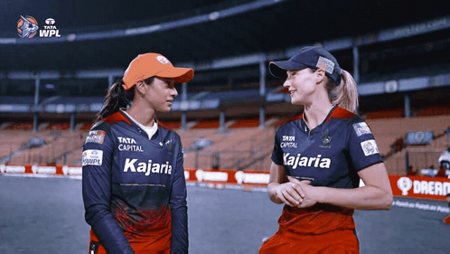 Two Women Wearing Kajaria Shirts Are Standing Next To Each Other On A Field GIF