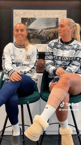 two women wearing sweaters that say coy 's are sitting on stools