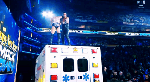 Two Wrestlers Are Standing On Top Of An Ambulance In Front Of A Sign That Says Mack Bank GIF
