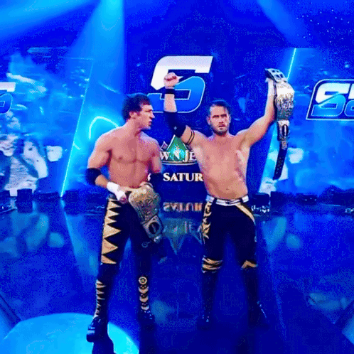 Two Wrestlers Holding Up Their Championship Belts In Front Of A Saturn Sign GIF