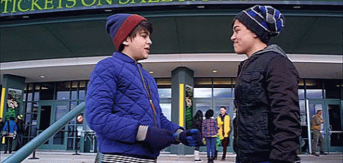 Two Young Boys Are Shaking Hands In Front Of A Building That Says Tickets On It GIF