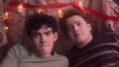 Two Young Men Are Laying Next To Each Other Under A Blanket With Christmas Lights . GIF