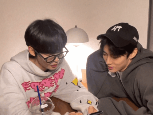 Two Young Men Are Sitting At A Table One Wearing A Hat That Says Ny GIF