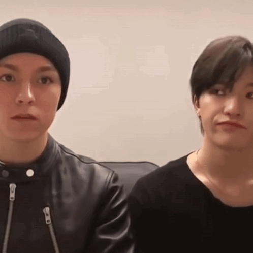 Two Young Men Are Sitting Next To Each Other And Looking At The Camera . One Of The Men Is Wearing A Beanie . GIF