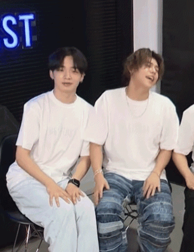 Two Young Men Are Sitting Next To Each Other In Front Of A Neon Sign That Says St GIF