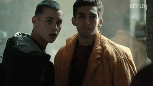 Two Young Men Are Standing Next To Each Other In A Dark Room . One Of The Men Is Wearing A Yellow Jacket . GIF