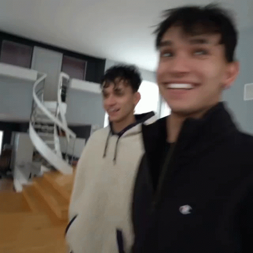 Two Young Men Are Standing Next To Each Other In A Room With A Spiral Staircase In The Background . GIF