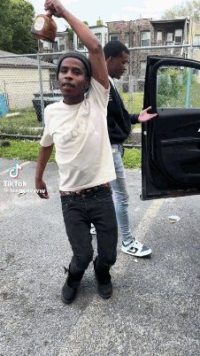 Two Young Men Are Standing Next To Each Other In Front Of A Car And One Is Holding A Bottle In His Hand . GIF