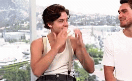 Two Young Men Are Standing Next To Each Other In Front Of A Window . One Of The Men Is Eating A Sandwich . GIF
