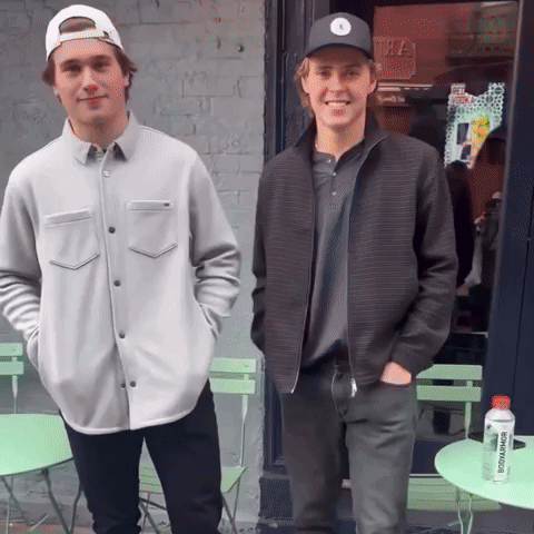 Two Young Men Standing In Front Of A Store With A Bottle Of Juice That Says ' Tropicana ' On It GIF