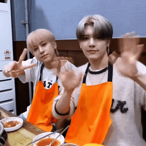 Two Young Men Wearing Aprons Are Sitting At A Table GIF
