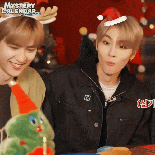 Two Young Men Wearing Santa Hats And Antlers Are Standing Next To A Christmas Tree And A Sign That Says Mystery Calendar GIF