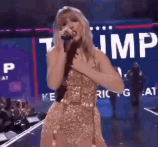Woman In Gold Dress Singing Into Microphone In Front Of Trump Sign GIF