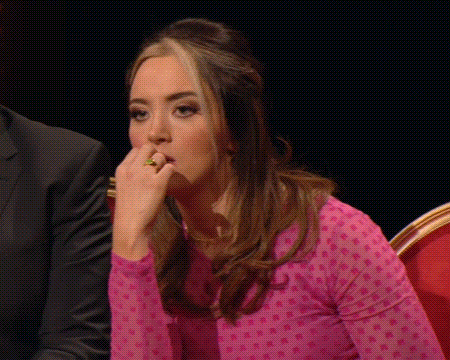 Woman In Pink Polka Dot Shirt Sitting With Hand To Mouth GIF