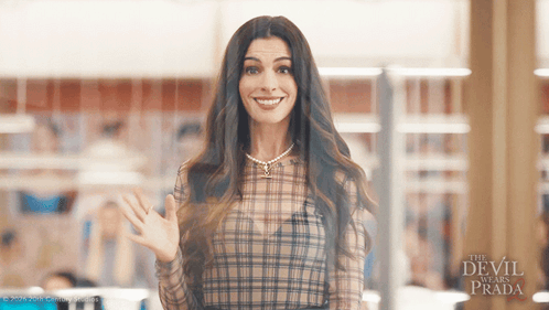 Woman In Plaid Shirt Smiling And Waving In Front Of The Devil Wears Prada Sign GIF