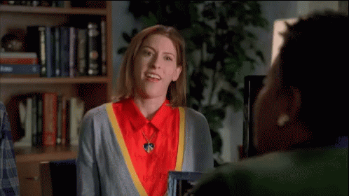 Woman in Red Shirt and Grey Sweater Waving at Another Woman in Front of Bookshelf