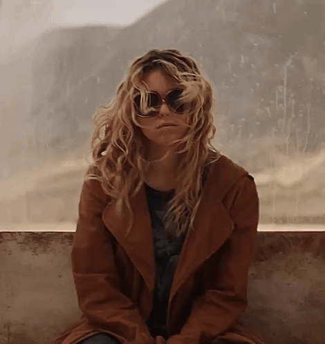 Woman In Sunglasses And Brown Coat Sitting On Wall GIF