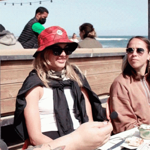 Woman in Sunglasses and Red Hat With Letter A Sits at Table Outside