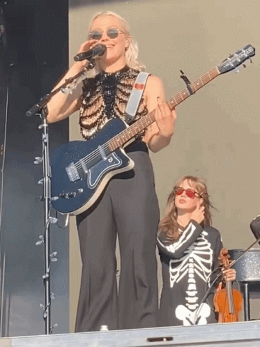 Woman Singing Into Microphone Holding Guitar Beside Girl in Skeleton Outfit