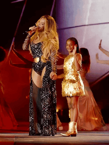 Woman Singing Into Microphone Next to Girl in Gold Dress