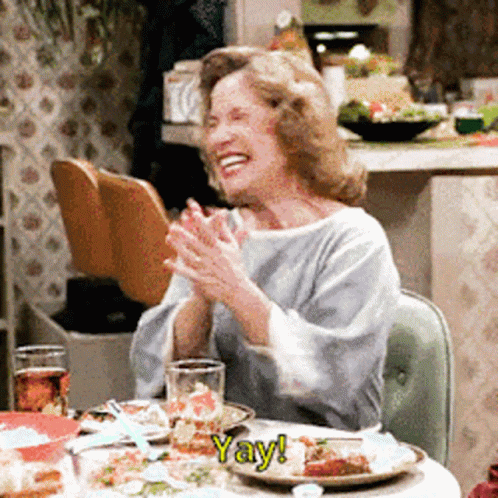Woman Sitting at Table With Food and Yay Sign