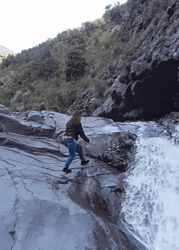 Woman Walking Down Rocky Cliff Near Waterfall