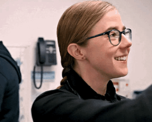 Woman Wearing Glasses and Black Shirt Smiling in Front of Telephone