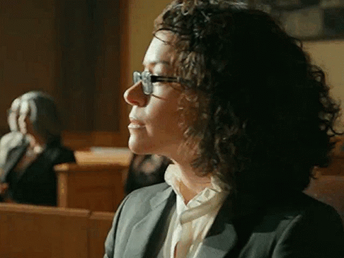 Woman With Curly Hair And Glasses Sitting In Courtroom GIF