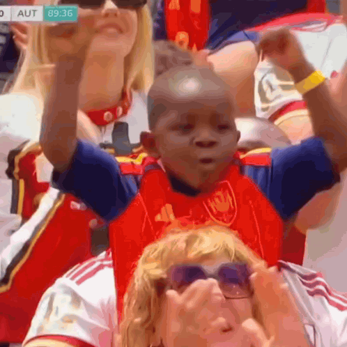 Young Boy in Red and Blue Adidas Shirt Sitting in Crowd