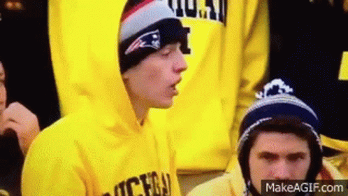 Young Man in Yellow Hoodie and Hat Sitting in Crowd