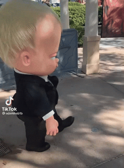 A Baby Doll In A Tuxedo Is Walking On A Sidewalk GIF