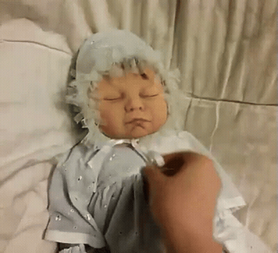 A Baby Doll Is Laying On A Bed With Its Eyes Closed And A Person Is Touching It . GIF