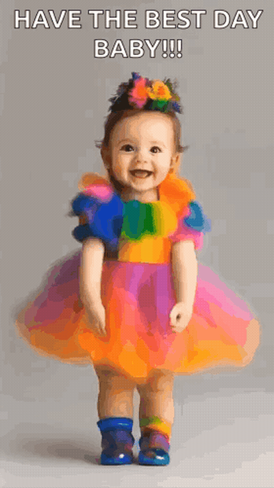 A Baby Girl In A Colorful Dress Is Standing And Smiling . GIF